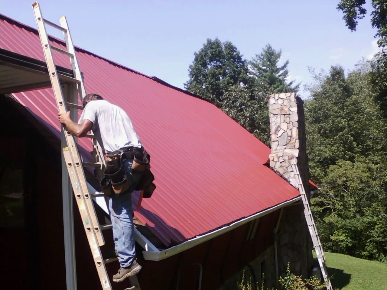 Roofer on a ladder beside a red metal roof and stone chimney — Soffit & Fascia Repair by Roofing Brothers in Grand Coteau